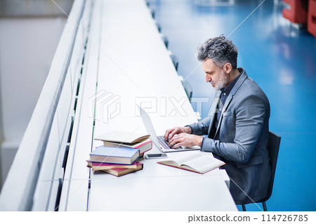Handsome businessman working on laptop, searching for informations in book. Traditional information source in modern office space. 114726785