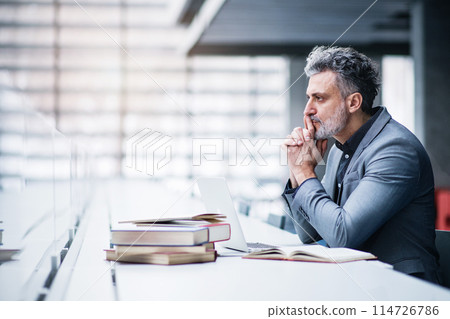 Handsome businessman working on laptop, searching for informations in book. Traditional information source in modern office space. 114726786