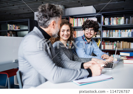 Handsome university professor helping students with final project, showing them research paper, book for thesis in library. 114726787