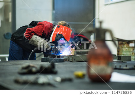 Beautiful blonde woman works as a welder in workshop, operating welding machine, wearing protective clothing and a welding mask. 114726826