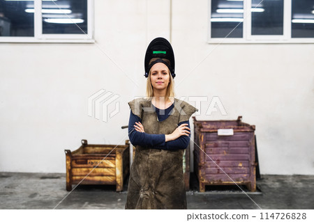 Beautiful blonde woman works as a welder in workshop, operating welding machine, wearing protective clothing and a welding mask. Beautiful blonde woman works as a welder in workshop, operating welding machine, wearing protective clothing and a welding mask. 114726828