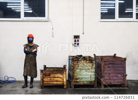 Beautiful blonde woman works as a welder in workshop, operating welding machine, wearing protective clothing and a welding mask. 114726831