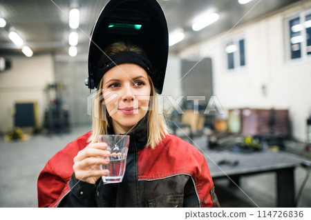 Beautiful blonde woman works as a welder in workshop, drinking water from glass. Wearing protective clothing and a welding mask. 114726836