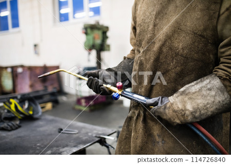 Beautiful blonde woman works as a welder in workshop, operating welding machine, wearing protective clothing and a welding mask. Beautiful blonde woman works as a welder in workshop, operating welding machine, wearing protective clothing and a welding mask. 114726840