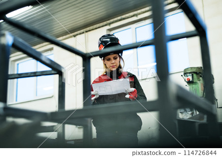 Beautiful blonde woman works as a welder in workshop, reading blueprints, wearing protective clothing and a welding mask. Beautiful blonde woman works as a welder in workshop, reading blueprints, wearing protective clothing and a welding mask. 114726844