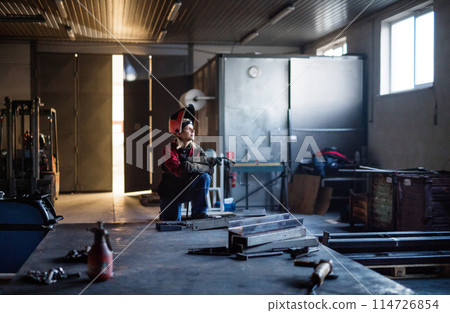 Beautiful blonde woman works as a welder in workshop, operating welding machine, wearing protective clothing and a welding mask. 114726854