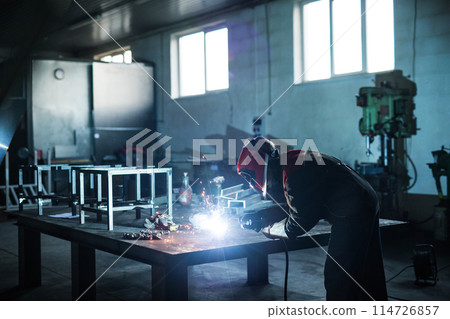 Beautiful blonde woman works as a welder in workshop, operating welding machine, wearing protective clothing and a welding mask. Beautiful blonde woman works as a welder in workshop, operating welding machine, wearing protective clothing and a welding mask. 114726857