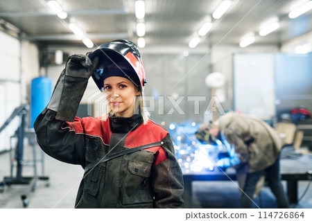 Beautiful blonde woman works as a welder in workshop, operating welding machine, wearing protective clothing and a welding mask. 114726864