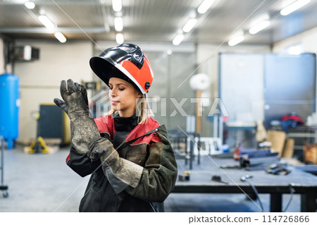 Beautiful blonde woman works as a welder in workshop, operating welding machine, wearing protective clothing and a welding mask. Beautiful blonde woman works as a welder in workshop, operating welding machine, wearing protective clothing and a welding mask. 114726866