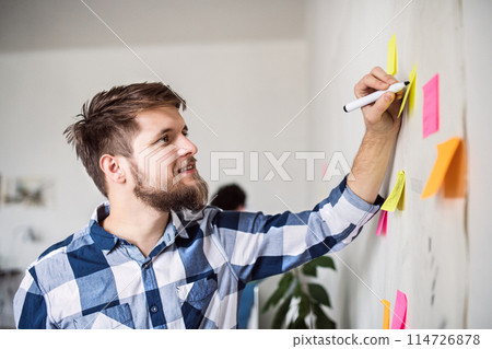 Man writing ideas on colorful sticky notes. Colleagues working on a new project together in a startup company office. Working in co-working space. 114726878