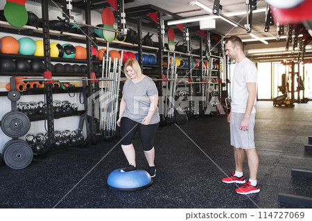 Overweight woman exercising on balance ball in gym. Personal trainer couching her and helping her. Overweight woman exercising on balance ball in gym. Personal trainer couching her and helping her. 114727069