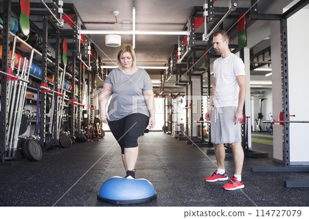 Overweight woman exercising on balance ball in gym. Personal trainer couching her and helping her. Overweight woman exercising on balance ball in gym. Personal trainer couching her and helping her. 114727079
