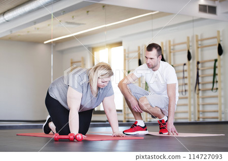 Overweight woman exercising on gym mat in gym. Personal trainer couching her and helping her. Overweight woman exercising on gym mat in gym. Personal trainer couching her and helping her. 114727093