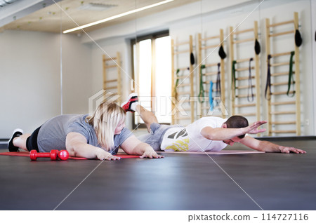 Overweight woman exercising on gym mat in gym. Personal trainer couching her and helping her. Overweight woman exercising on gym mat in gym. Personal trainer couching her and helping her. 114727116