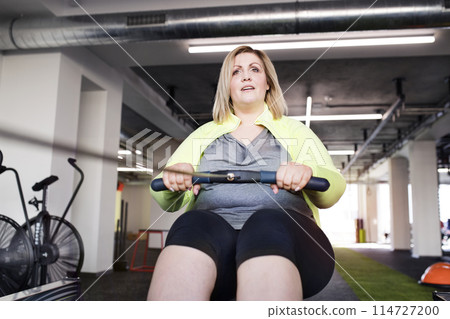 Overweight woman exercising on rowing machine. Routine workout for physical and mental health Overweight woman exercising on rowing machine. Routine workout for physical and mental health 114727200