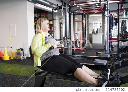 Overweight woman exercising on rowing machine. Routine workout for physical and mental health Overweight woman exercising on rowing machine. Routine workout for physical and mental health 114727202