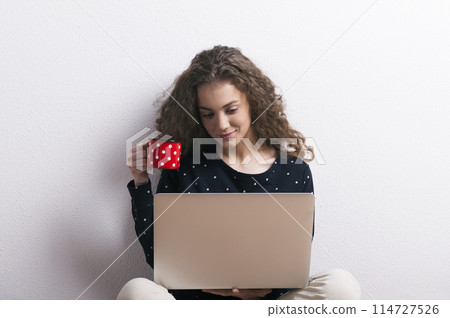 Portrait of gorgeous teenage girl sitting, typing on notebook, drinking coffee. Studio shot, white background with copy space Portrait of gorgeous teenage girl sitting, typing on notebook, drinking coffee. Studio shot, white background with copy space 114727526