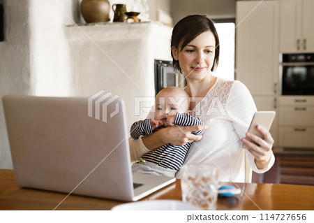 Mother with small baby working from homeoffice, typing on laptop. Mom paying bills online, holding small baby. 114727656