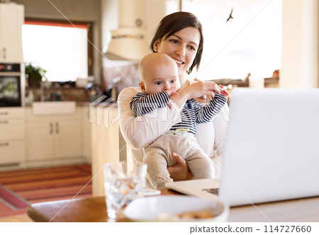 Mother with small baby working from homeoffice, typing on laptop. Mom paying bills online, holding small baby. 114727660
