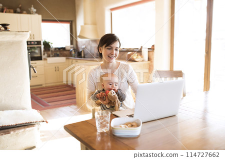Mother with small baby working from homeoffice, typing on laptop. Mom paying bills online, holding small baby. 114727662