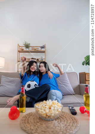 Lesbian couple cheering for Euro football at home with popcorn and drinks. Concept of sports enthusiasm and LGBTQ pride 114728957