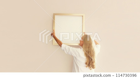 Woman decorating interior, hanging white blank photo frame, wooden picture on wall in new house Woman decorating interior, hanging white blank photo frame, wooden picture on wall in new house 114730396