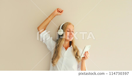 Happy cheerful woman listening to music with mobile phone in headphones having fun dancing Happy cheerful woman listening to music with mobile phone in headphones having fun dancing 114730397