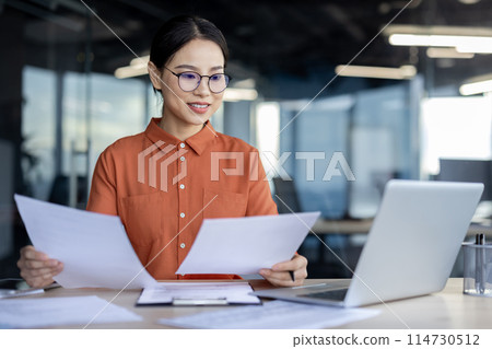 Asian businesswoman analyzing documents at desk in a modern office while working on a laptop. Professional, focused, and productive work environment. 114730512
