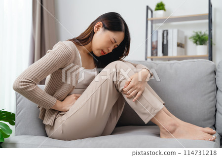 The woman with a stomachache is holding her stomach in pain, sitting on the sofa in the living room. 114731218