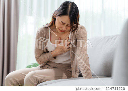 A woman is sitting on a couch with her hand holding on her chest, suffering from a heart attack. 114731220