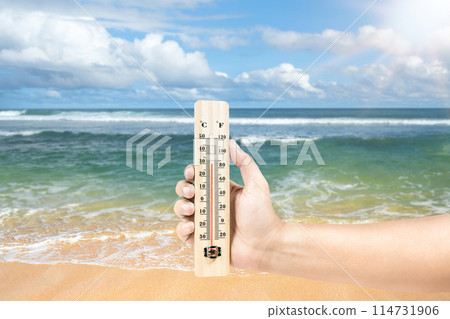 High temperature weather on summer seasons. Image of human hand holding thermometer on a beach with sunbeam. 114731906
