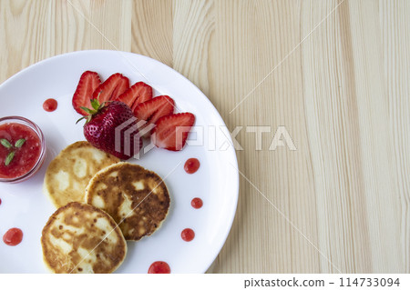 Three pancakes with cheese and strawberries and sauce on a white dish against wood background. Delicious cheese pancake and strawberries for the breakfast or lunch. Tasty meal with berries. Three pancakes with cheese and strawberries and sauce on a white dish against wood background. Delicious cheese pancake and strawberries for the breakfast or lunch. Tasty meal with berries. 114733094