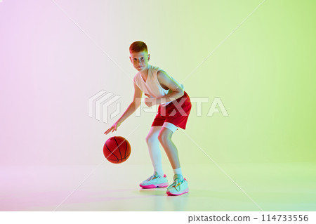 Focused boy, basketball player training, playing, dribbling ball against gradient background in neon light 114733556