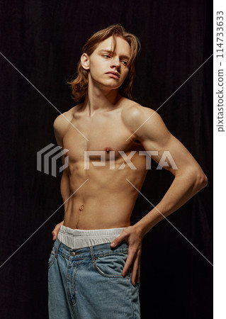 Handsome young man standing shirtless, with muscular, relief strong body posing in jeans against black background. 114733633