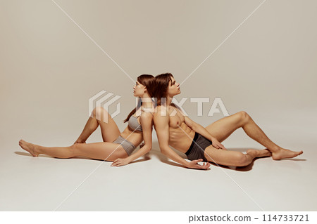 Athletic models, young woman and handsome man sitting on floor in inner wear against warm grey background Athletic models, young woman and handsome man sitting on floor in inner wear against warm grey background 114733721