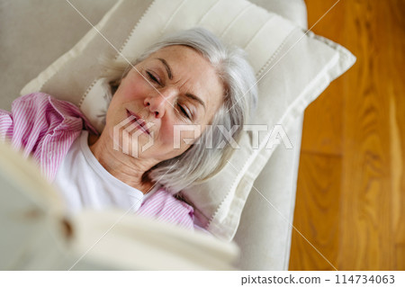 Beautiful mature woman reading book, feeling cozy and happy. Weekend activity for older woman, relaxing at home. Top view Beautiful mature woman reading book, feeling cozy and happy. Weekend activity for older woman, relaxing at home. Top view 114734063