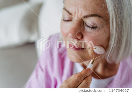 Skincare for mature woman. Older woman using facial roller for blood circulation, after serum. 114734067