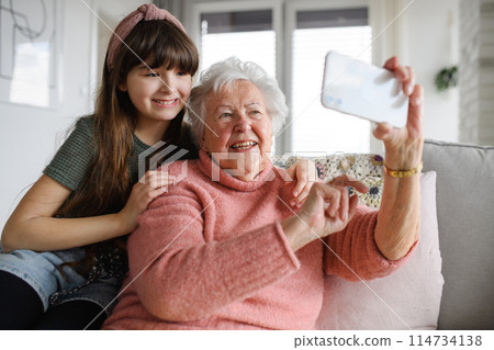 Grandmother with cute girl taking selfie with smartphone. Portrait of an elderly woman spending time with granddaughter. Grandmother with cute girl taking selfie with smartphone. Portrait of an elderly woman spending time with granddaughter. 114734138