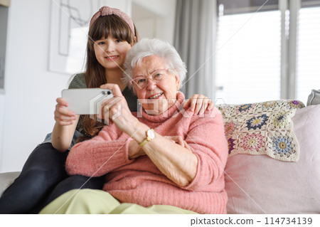 Grandmother with cute girl taking selfie with smartphone. Portrait of an elderly woman spending time with granddaughter. Grandmother with cute girl taking selfie with smartphone. Portrait of an elderly woman spending time with granddaughter. 114734139