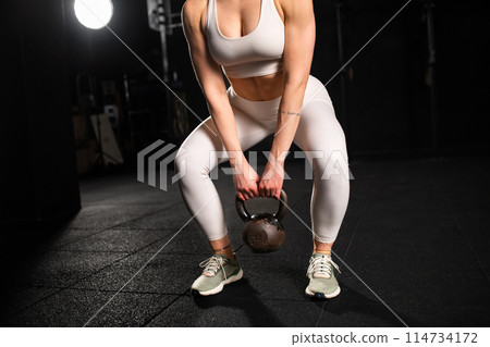 Woman performing kettlebell swings, holding kettlebell in both hands. Routine workout for physical and mental health. Woman performing kettlebell swings, holding kettlebell in both hands. Routine workout for physical and mental health. 114734172