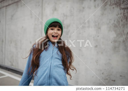 Outdoor portrait of cute young girl with beanie hat. Girl with long hair standing on playground in the city. 114734271