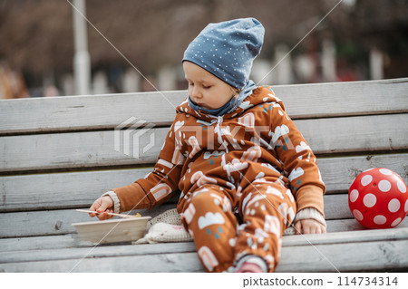 Cute toddler girl eating lunch in park, sitting on bench. Picnic in park during cold spring day. 114734314