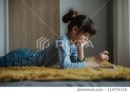 Cute girl lying on belly, scrolling on smartphone. Using social media. Side view. Cute girl lying on belly, scrolling on smartphone. Using social media. Side view. 114734316