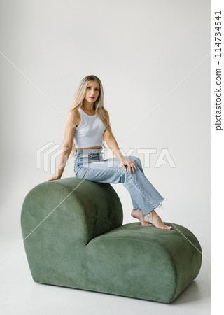 Young blonde woman is sitting on a green modern sofa. She's wearing a white T-shirt and blue jeans 114734541