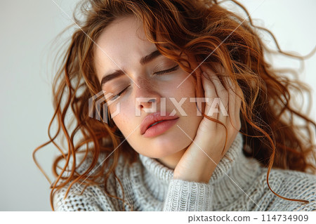 Young red haired woman in a white sweater rests her head on her hand. Does she have a headache or toothache 114734909