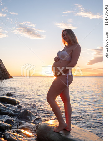 Pregnant woman in bikini posing on rocky beach at sunrise with mountain view Pregnant woman in bikini posing on rocky beach at sunrise with mountain view 114735063