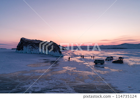 Aerial winter landscape of frozen lake Baikal. Groups of tourists got off the cars and walking around the rocky island in lake Baikal, walking on the crystal clear ice at sunrise. Famous tourist spot 114735124
