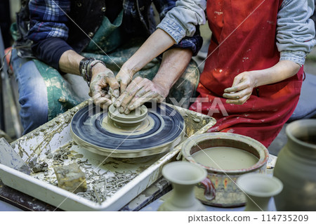 Potter teaching child to work on potter wheel, how to sculp clay pot. Closeup of dirty hands Potter teaching child to work on potter wheel, how to sculp clay pot. Closeup of dirty hands 114735209