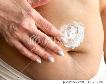 Close up of womans hand with white manicured nails applying cream to her stomach. Highlights skincare, body care, and personal hygiene, focusing on moisturizing and self care routines. Close up of womans hand with white manicured nails applying cream to her stomach. Highlights skincare, body care, and personal hygiene, focusing on moisturizing and self care routines. 114735491