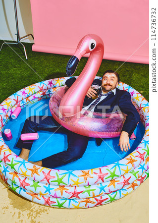 Young office worker in formal suit relaxing in kiddies swimming pool on flamingo inflatable-ring and drinks beer. Young office worker in formal suit relaxing in kiddies swimming pool on flamingo inflatable-ring and drinks beer. 114736732